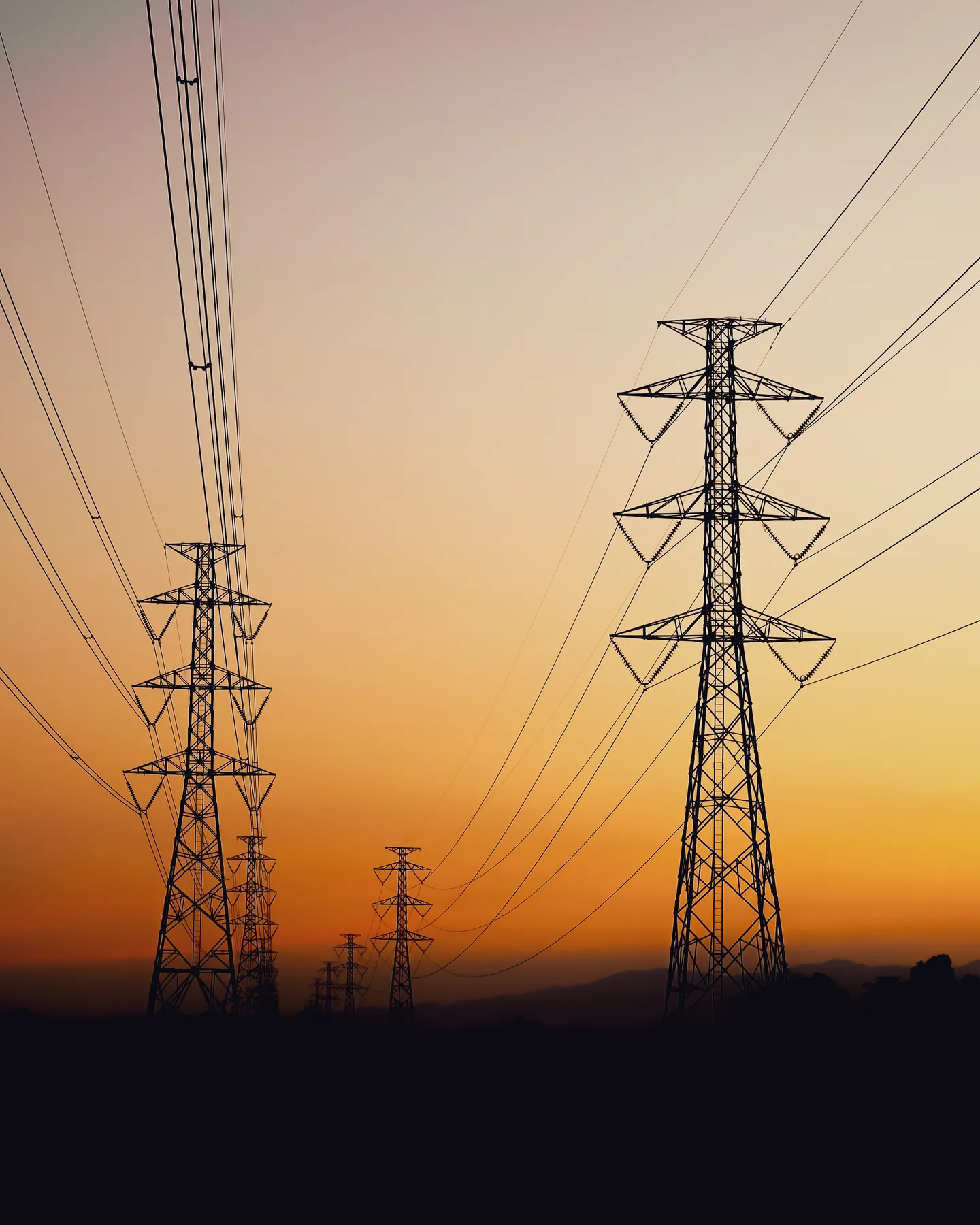 High-voltage lattice transmission towers carrying conductor across open country at sunset.