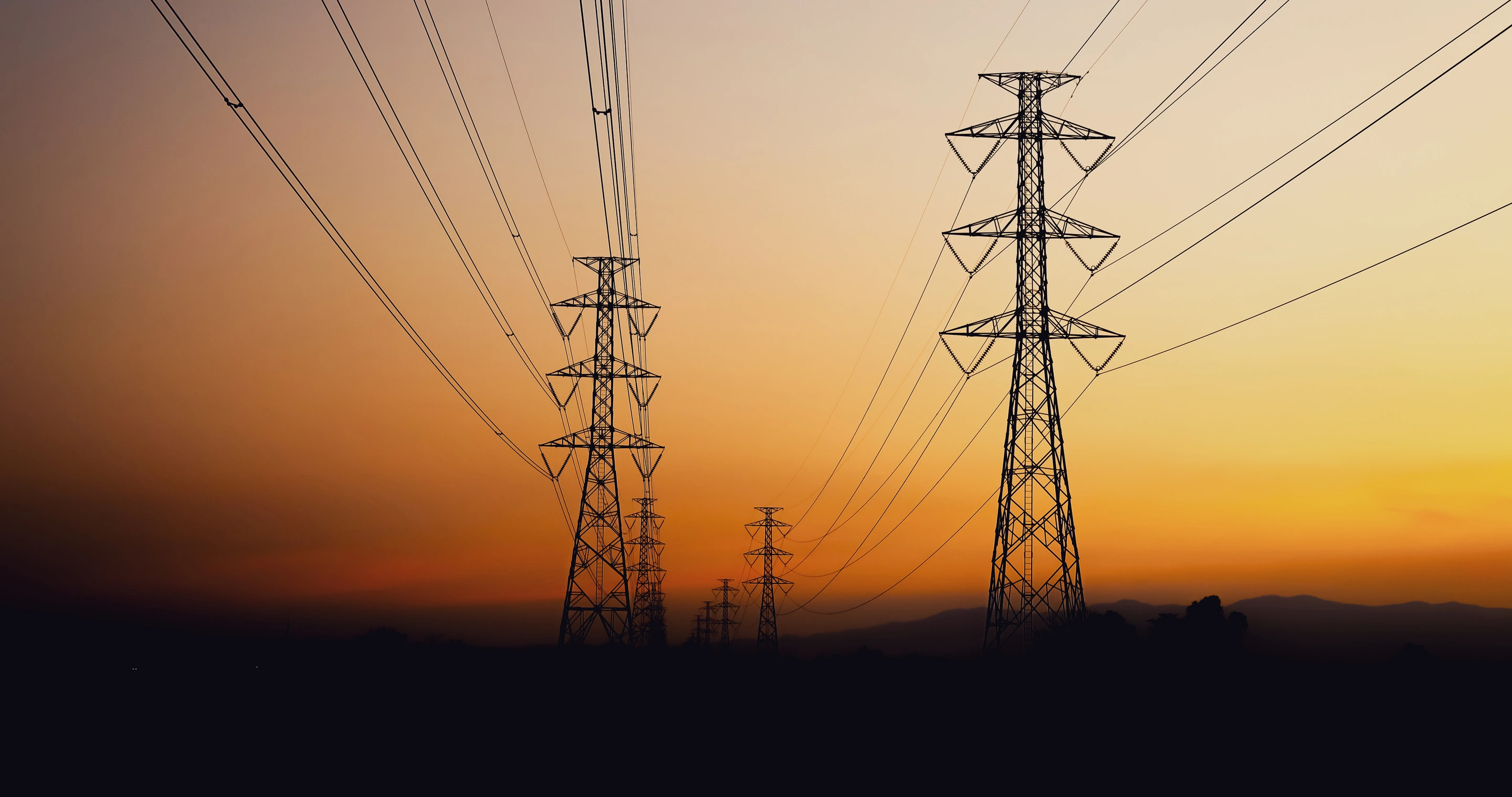 High-voltage lattice transmission towers carrying conductor across open country at sunset.